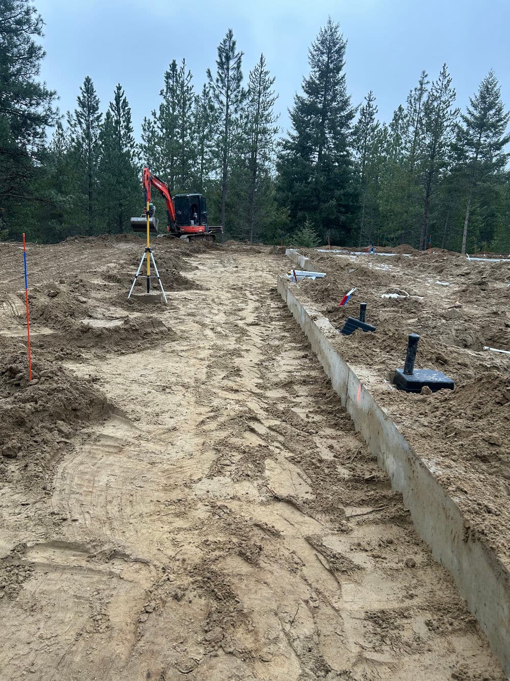 Construction site showing a prepared foundation, surveying equipment, and excavation machinery.
